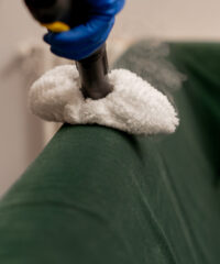 close up in an apartment cleaner in yellow apron does a steam cleaning of a green sofa with professional equipment